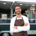 Smiling young ethnic male waiter in apron standing with arms folded near modern food truck and looking at camera contentedly